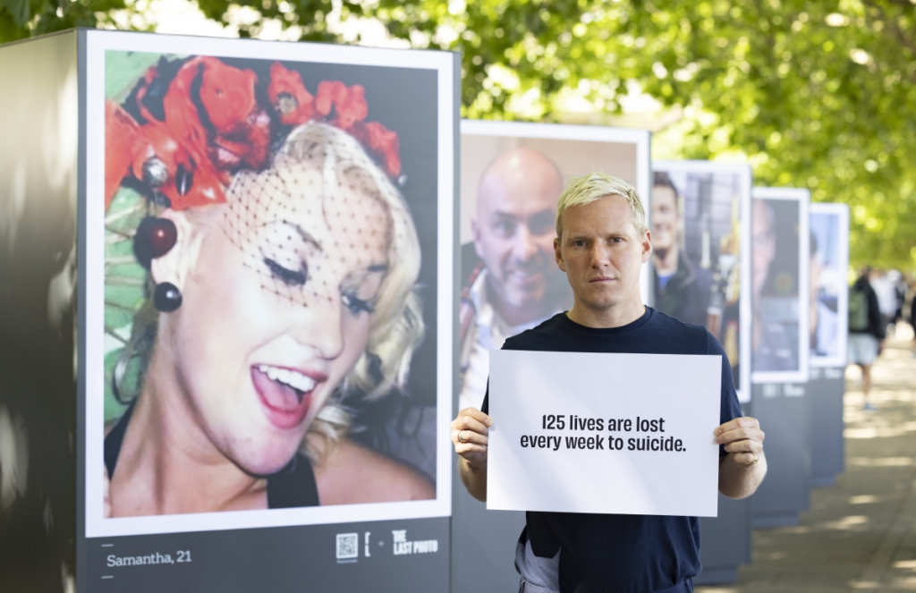 a man standing in front of a row of "The Last Photo" images holding a sign that reads "123 lives are lost every week to suicide".